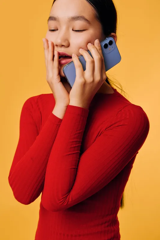 Young woman in red sweater holding smartphone near face with closed eyes, expressing emotion or deep thought on yellow background. Female, technology and communication concept.