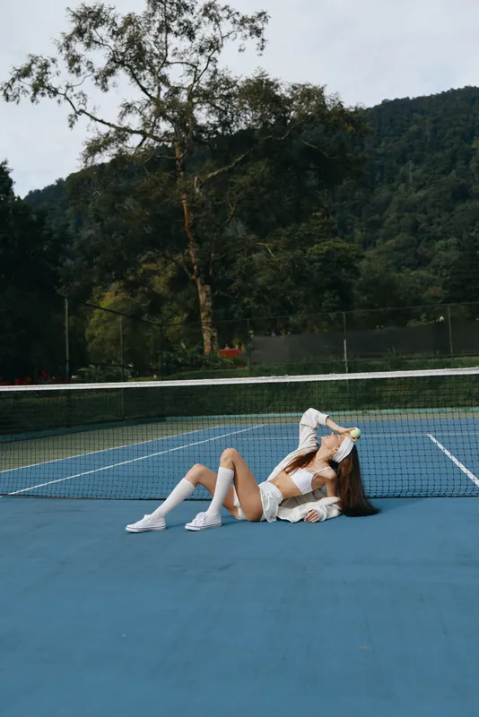 Young female tennis player relaxing on the court, showcasing sporty attire, emotions of joy and tranquility, amidst a vibrant, natural background