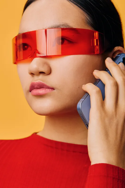 Young woman with futuristic red visor glasses talking on smartphone in closeup portrait against yellow background. Modern fashion, technology, communication, accessories and trendy style.