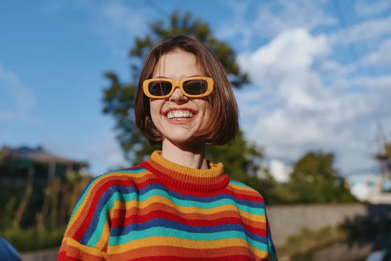 Young woman in a rainbow sweater and yellow sunglasses smiling outdoors under a sunny blue sky. Short hair bob, early 20s female model in casual outfit, joyful lifestyle portrait.