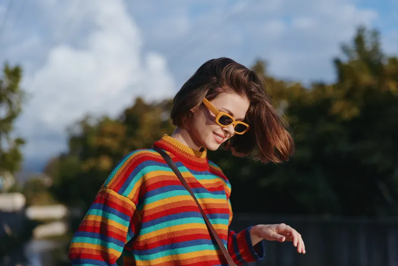 Woman in a vibrant striped sweater and yellow sunglasses walking outdoors, smiling and relaxed, casual outfit with shoulder bag in urban park lifestyle fashion travel scene