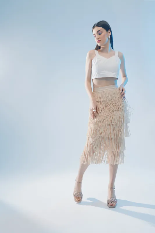 Young woman in a white crop top and beige fringe skirt poses elegantly on a light background. Concept of beauty, fashion, and modern style with soft lighting.
