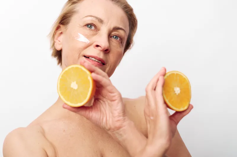 Mature woman with natural skin holding two orange halves near face with cream on cheek. Concept of skincare, healthy lifestyle, natural beauty, vitamin c, and self-care routine.