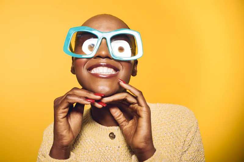 Smiling black woman with dark skin wearing large blue glasses and beige sweater poses against yellow background. Concept for joy, fashion, and confident beauty portrait.