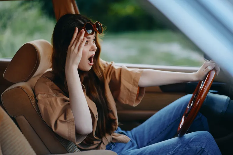 Shocked female driver in casual outfit, expressing surprise and frustration in a car during a sunny day, green background adds depth and contrast.