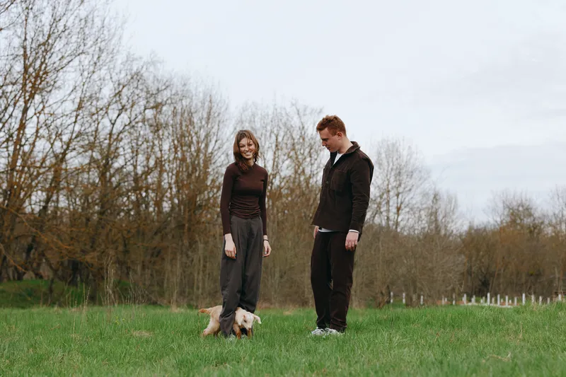 A couple outdoors with a small dog in a grassy park, casual outfits, joyful mood, relaxed interaction, natural light, candid moment, lifestyle scene, affectionate companionship, daytime stroll,