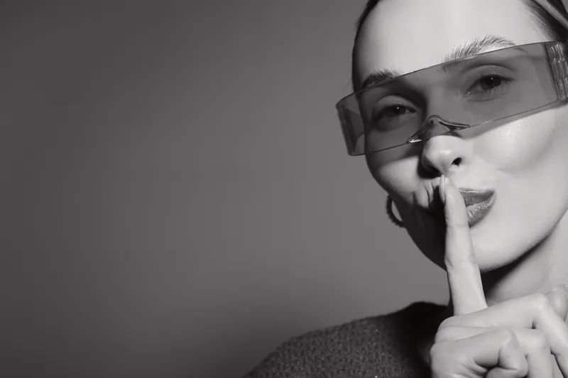Close-up of a young Caucasian woman with fair skin wearing transparent glasses making shushing gesture. Concept for silence, secrecy, or quiet communication.