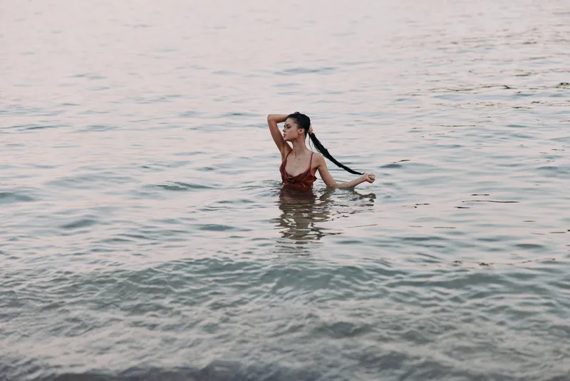 A lone swimmer stands waist deep in calm water, hair tied back, wearing a red swimsuit, with gentle ripples and soft light across the scene.