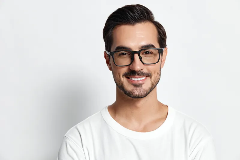 Smiling happy cheerful young man in casual white t-shirt wearing glasses looks at the camera, showcasing a warm and friendly demeanor against a plain background.