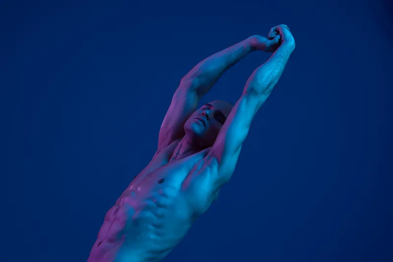 Artistic male figure stretching against a vibrant blue backdrop, showcasing muscle definition and unique lighting. Evokes strength and fluidity in motion.