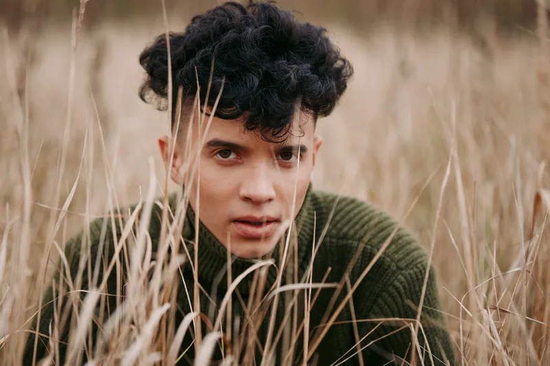 Close portrait of a young person crouching in dried tall grass, conveying raw authenticity and quiet reliability through natural expression, textured sweater, and warm earthy tones.