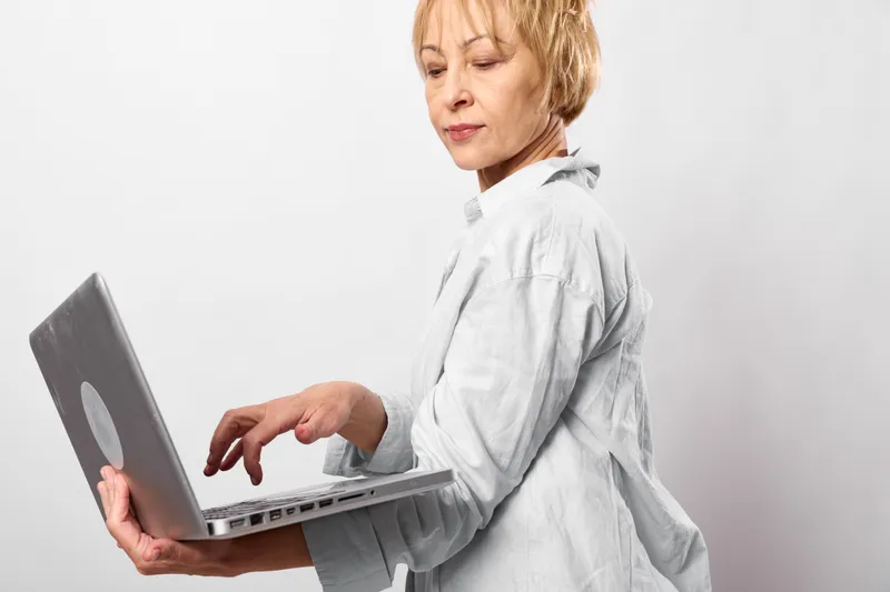 Woman with laptop typing, in white coat, elderly medical professional, healthcare clinic work, focused serious face, holding computer, indoor studio background, remote consultation