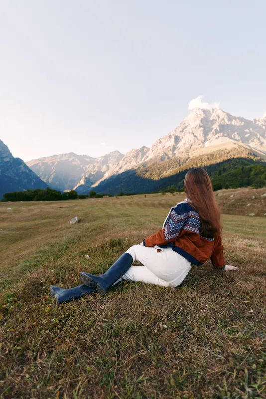 Woman sitting on meadow overlooking mountains and landscape, cozy sweater and boots in nature, relaxed outdoor scene for travel, hiking, solitude and scenic countryside view.