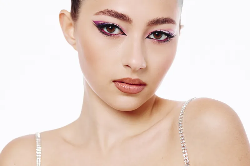 Closeup of a young woman with bold bi-color eyeliner looking at the camera in a white studio. Clear skin and glitter straps reflect modern beauty, fashion, and creativity concepts.