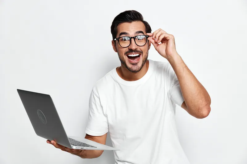Excited smiling man wearing glasses holds laptop and looks at camera with joyful expression on white background. Modern technology lifestyle concept.