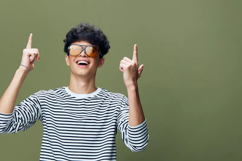 smiling man wearing striped shirt and stylish sunglasses pointing upwards with both hands on green background, joyful young adult with curly hair expressing excitement