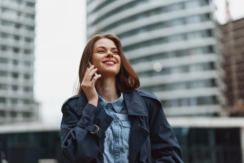 A cheerful young woman with brunette hair chats on her phone in an urban street setting, wearing a denim shirt and a dark coat, smiling.