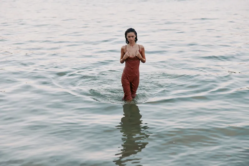 A solitary figure stands waist deep in calm water, wearing reddish overalls, contemplative pose, gentle ripples surround, soft light, tranquil scene, natural reflection, minimal and serene atmosphere.