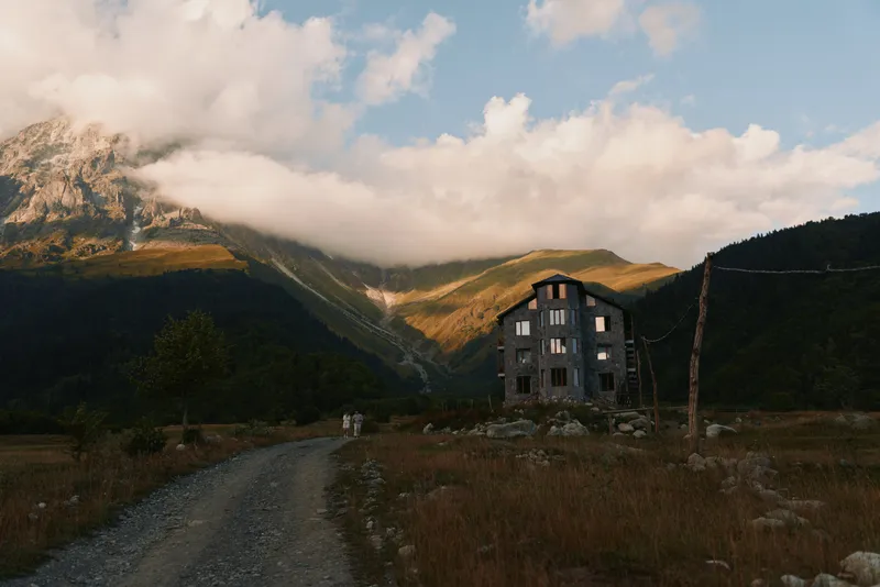 A weathered mountain house sits beside a dirt road as clouds roll over rugged peaks, conveying solitude, resilience, and quiet beauty in a remote landscape.