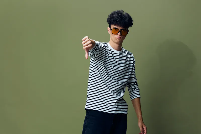 Young man wearing sunglasses and striped shirt showing thumbs down gesture expressing disapproval against green background in casual style.