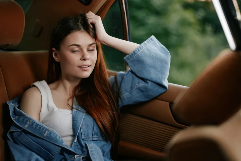 Young woman with long red hair wearing a denim jacket, relaxing in a car, conveying a sense of tranquility and introspection in a natural green setting.
