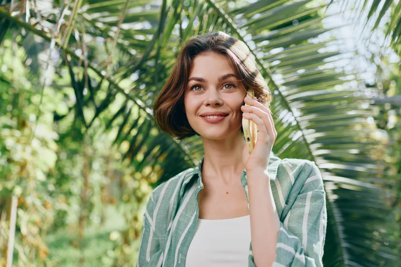 Portrait of a smiling woman standing outdoors among lush tropical greenery, sunlight on her face, casual cardigan and white top.