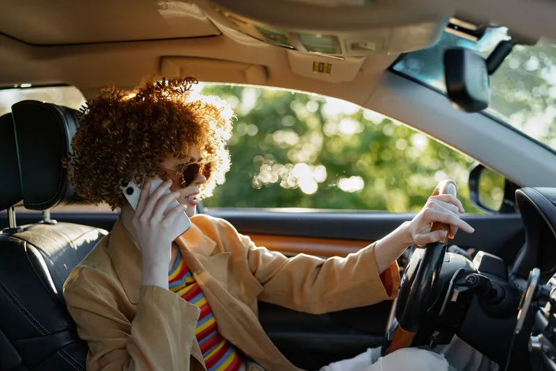 Cheerful woman driving car, talking on phone, curly hair, retro color vibe, sunlit interior, casual blazer, striped shirt visible, candid moment, natural surroundings