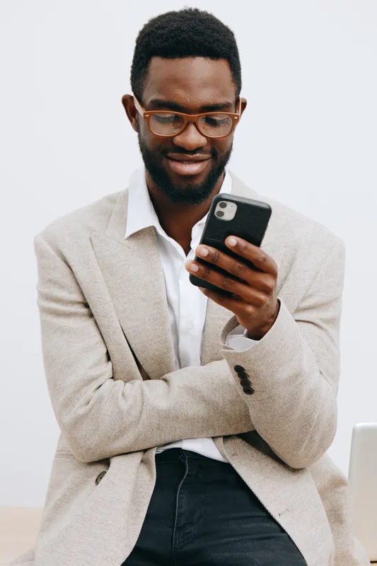 Smiling man with dark skin and glasses using smartphone on plain white background, casual business attire, vertical shot, studio portrait, modern technology, lifestyle concept, professional vibe