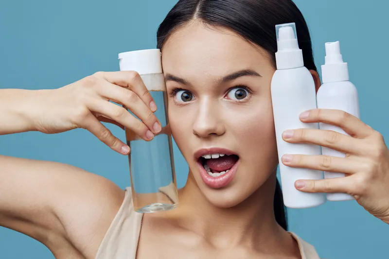 Young woman excitedly showcasing skincare products with a playful expression, bright background, emphasizing beauty and self care routines