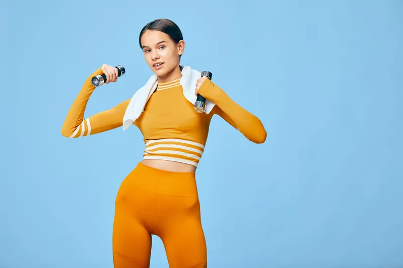 Woman fitness in orange activewear lifts dumbbells with a towel draped over her shoulders, posing confidently against a solid blue backdrop for a high-energy workout moment and strong athletic