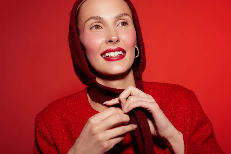 Smiling woman in red sweater and hood poses against red background. She looks happy and confident with natural makeup and hoop earrings. Concept for fashion, winter, and beauty.