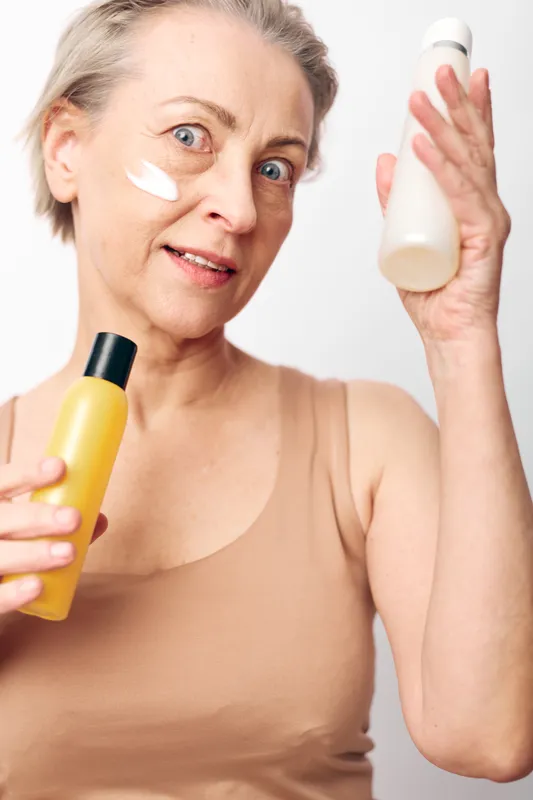 Mature woman with short hair applies white facial cream on cheek holding two cosmetic bottles. Studio portrait on white background. Concept of skincare, beauty routine, and self care.