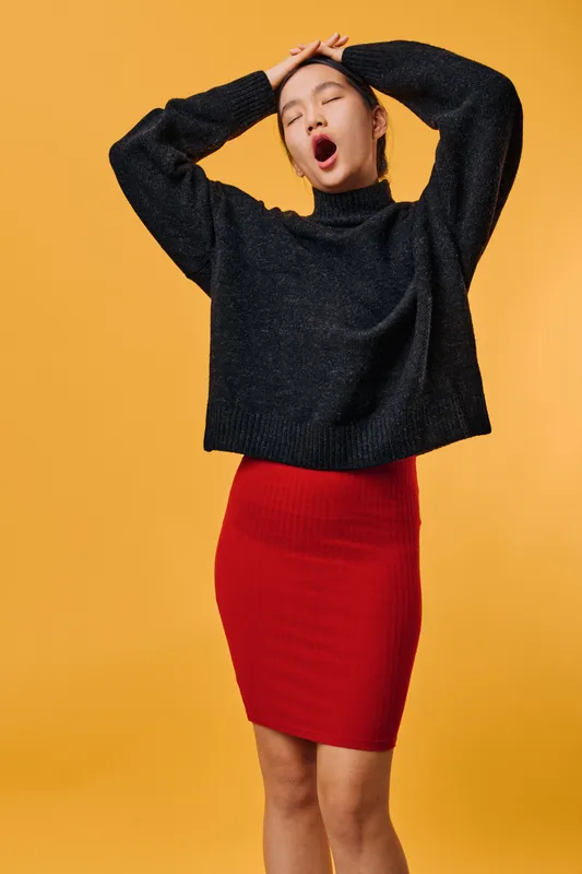 Young woman yawning and stretching with hands on head wearing black sweater and red skirt against yellow background. Casual style, tired expression, isolated studio portrait with copy space.