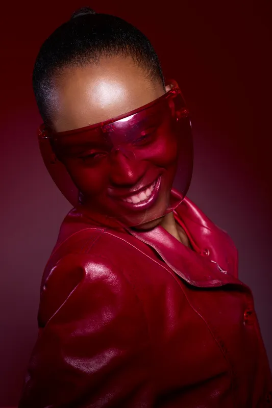 Portrait of a smiling dark skinned African woman wearing a red leather jacket and red transparent visor. Studio lighting highlights her radiant skin and joyful expression. Fashion concept.