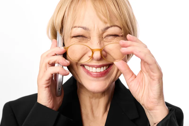 Cheerful smiling mature woman, blonde hair, adjusting round eyeglasses with fingertips, holding a phone near ear, close up portrait, bright studio light, clean white background