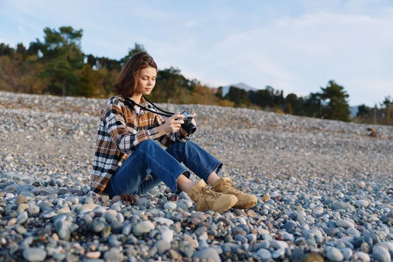 Woman with camera sits on a rocky beach, dressed in a plaid shirt and boots, focused on photography in a calm outdoor setting with distant trees and blue sky, capturing a quiet moment