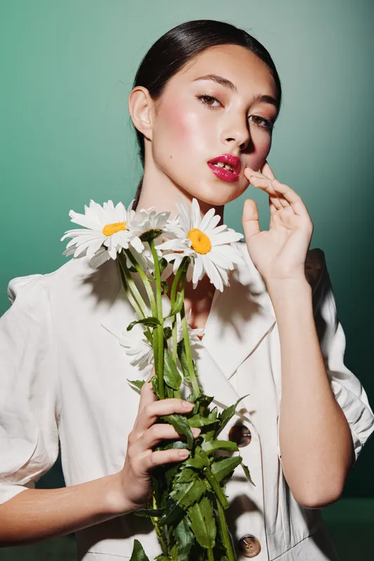 Fashionable young woman with glossy red lips holding white daisies in a white blazer.Editor styled creativity, bright studio with soft lighting.Flat makeup concept with natural floral mood.