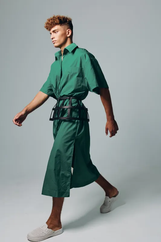 Young man walking in green oversized shirt and matching wide pants with black harness belt and white slip on shoes in studio with neutral background and casual style.
