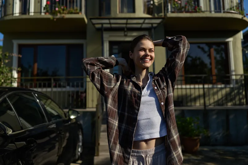 Smiling woman outdoors in casual plaid shirt, hands run through hair. Sunny daytime setting highlights carefree lifestyle and confident mood