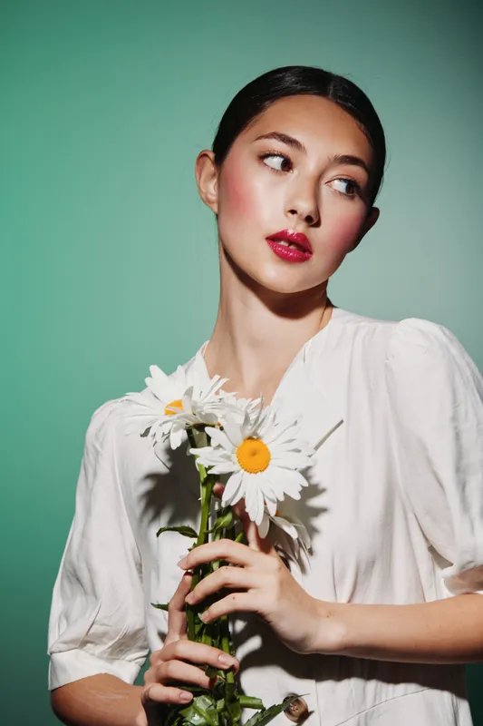 Young woman with glossy red lips and soft makeup holds white daisies against a muted green background wear a stylish white blouse. Concept of beauty fashion flower elegance.