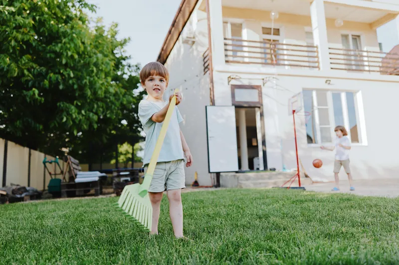 Playful boy holding a rake in a backyard, enjoying a sunny day outdoors while a girl plays basketball nearby. A perfect scene of childhood happiness and fun.
