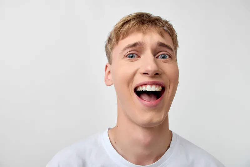 Surprise, excited man expression with joyful amazed closeup face, wide open mouth, studio lighting, white shirt, casual emotion and energy, vibrant candid moment, cheerful gaze and camera frame with