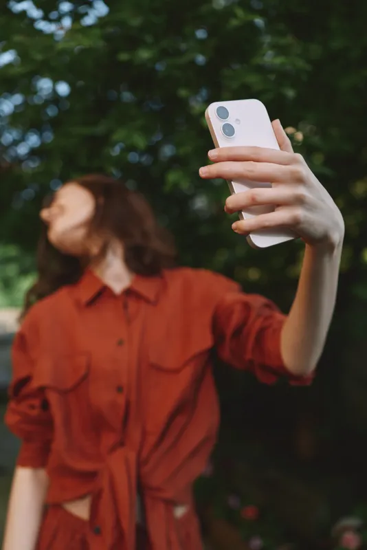 Young woman with brunette hair wears an orange shirt and takes a candid selfie in a leafy park, enjoying a spontaneous outdoor moment with a playful mood