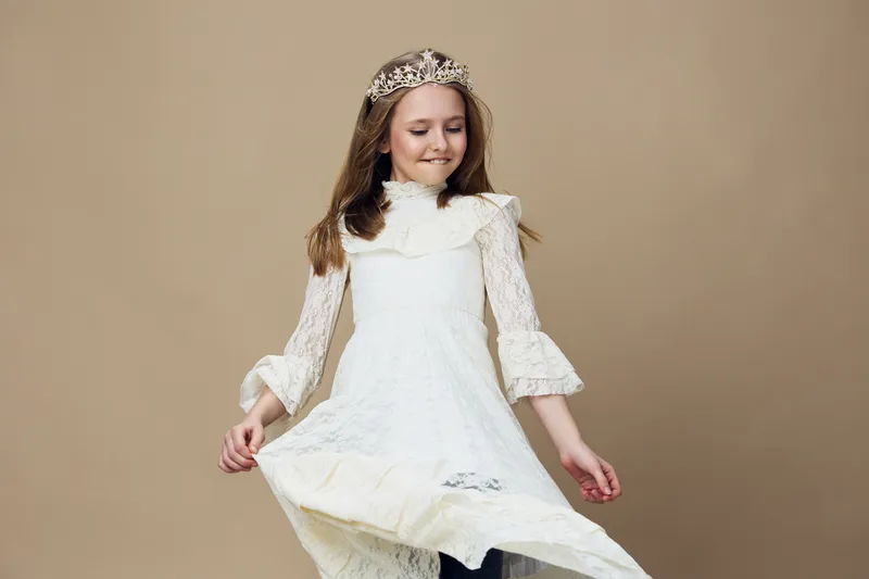 Delightful young girl wearing a lace dress and crown, joyfully twirling with a natural smile against a soft beige background, embodying innocence and happiness