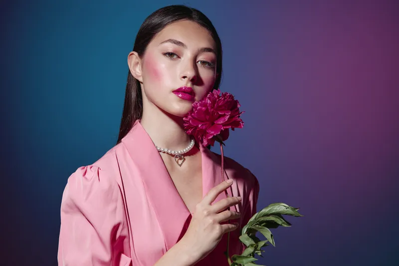 Young woman wearing a pink coat holds a deep pink flower near her face. Her makeup is dramatic with bright lipstick and highlighter emphasizing beauty and fashion style.