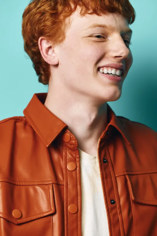 Smiling young man with red hair wearing brown leather jacket and white shirt against blue background in bright studio portrait. Happy casual fashion and positive expression.