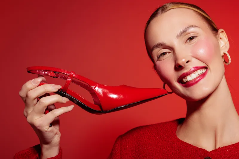 Smiling woman with red lipstick holds shiny red high heel shoe near face on red background. Concept of fashion, beauty, style, and glamour with vibrant color contrast and joyful mood.