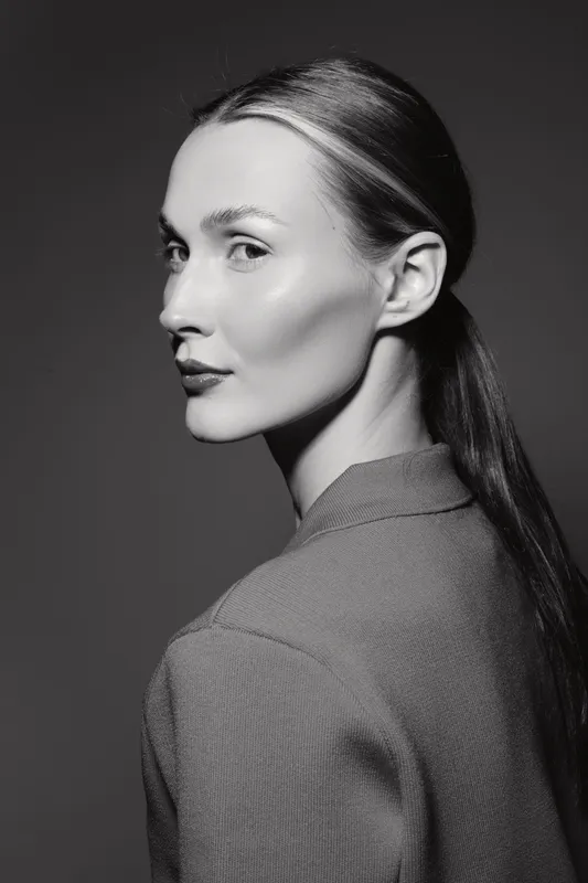 Profile portrait of a young Caucasian woman with fair skin and sleek hair, wearing a textured jacket. Studio black and white photography with soft lighting and contrast.