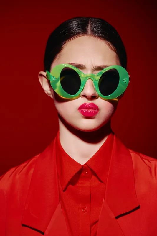 Young woman with slick hair wears bold green sunglasses and bright red lipstick against crimson backdrop. High fashion style contrasts vivid colors creating striking beauty concept.
