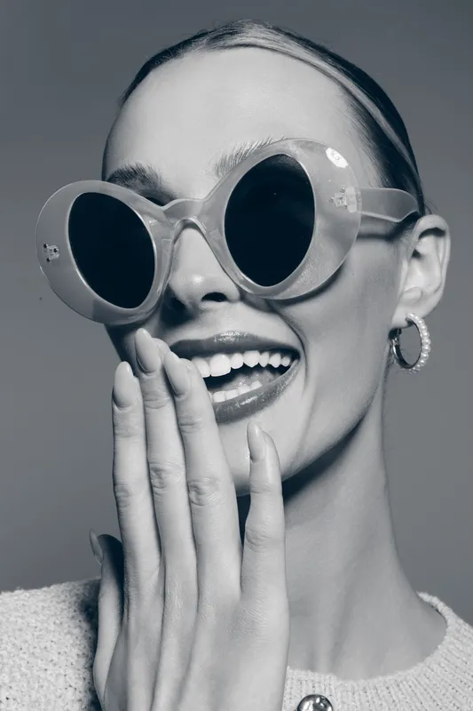 Happy white woman with light skin wearing large sunglasses laughs covering mouth with hand. Studio portrait in black and white.
